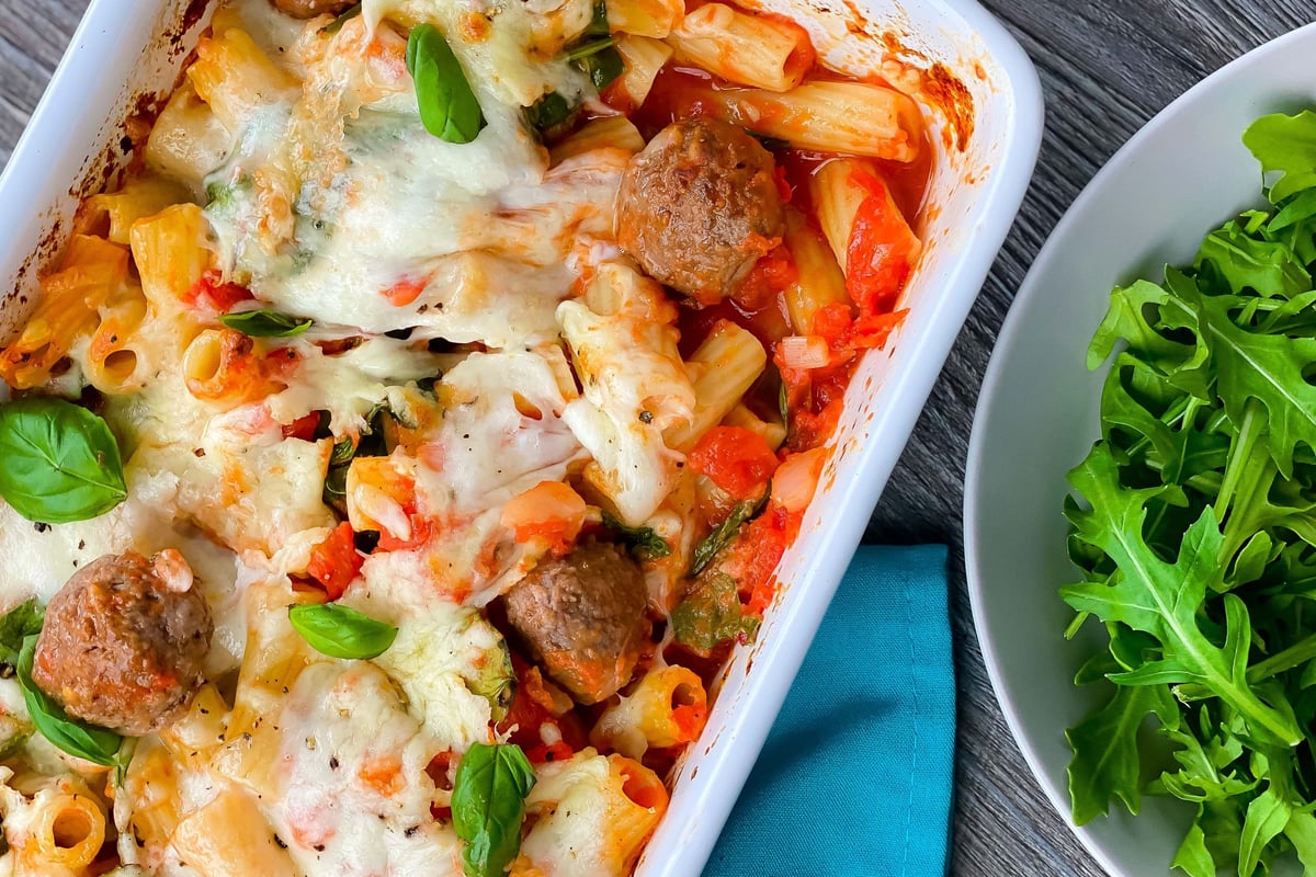 Baked pasta with meatballs, melted cheese and basil in white dish beside rocket salad
