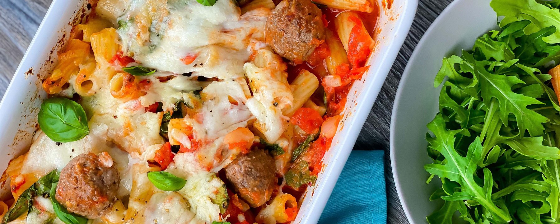 Baked pasta with meatballs, melted cheese and basil in white dish beside rocket salad