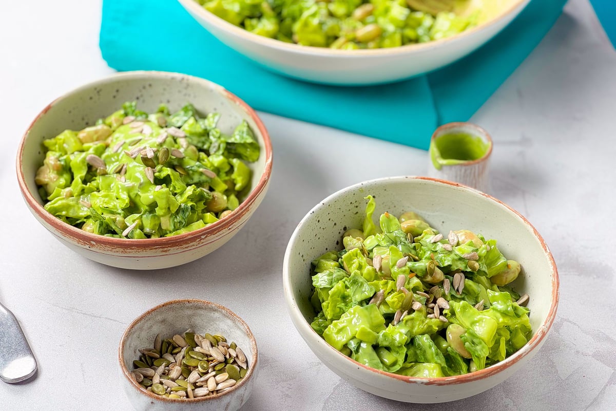 Two bowls of green salad with seeds and a small bowl of mixed seeds on a white surface