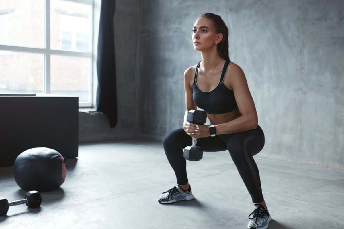 Woman in sportswear doing squats holding a dumbbell in a gym room.