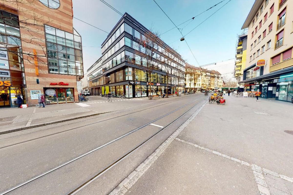 PureGym Basel Clara Huus: Eine urbane Strasse mit Geschäften, Strassenbahngleisen und PureGym-Beschilderung in einem Fenster gegenüber.