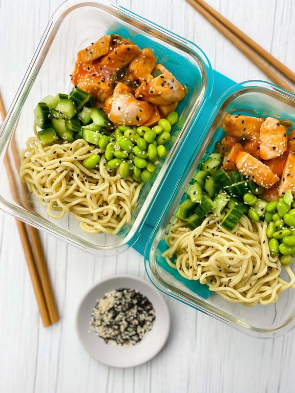 Two glass containers with noodles, diced cucumber, edamame, and sesame chicken pieces