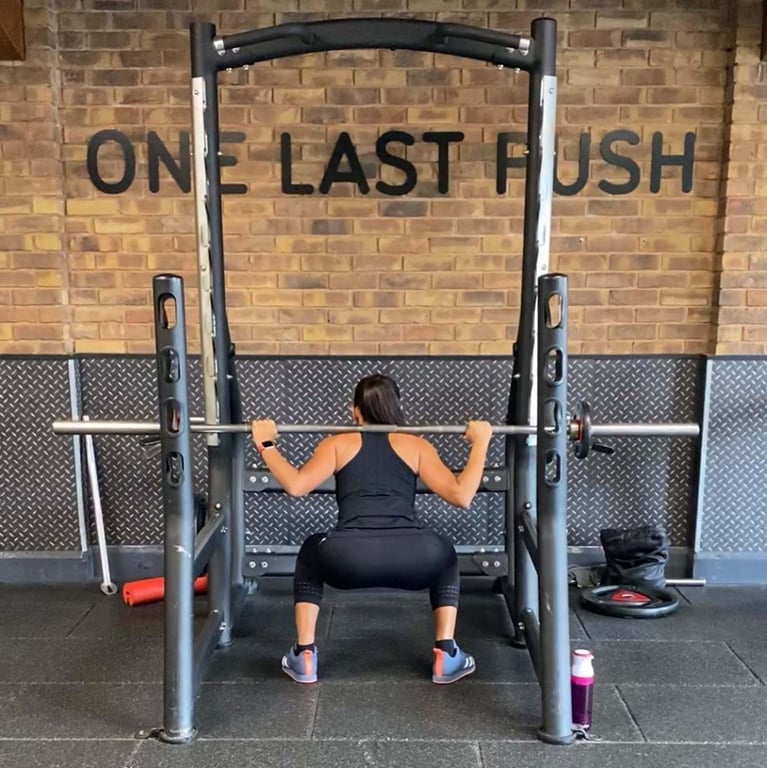 Woman squatting with barbell on shoulders inside gym rack with "One Last Push" sign behind