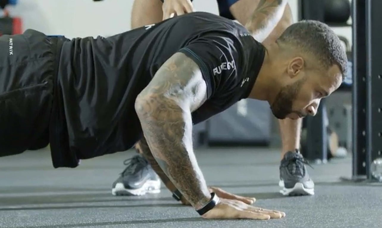 Man with tattoos doing a push-up in gym with another person standing behind him