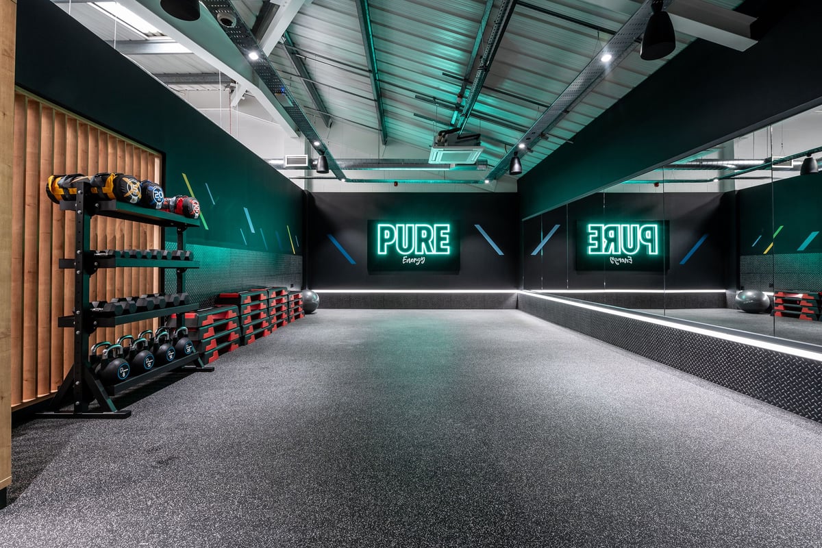 PureGym Guiseley: Dumbbell racks, weight plates, teal walls, rubberized flooring, neon 'PURE' signage.