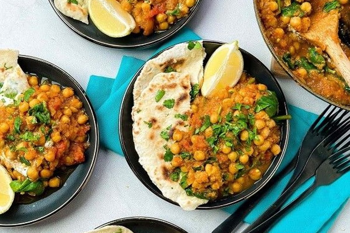 Four plates of chickpea curry with flatbread and lemon wedges next to a pan of curry