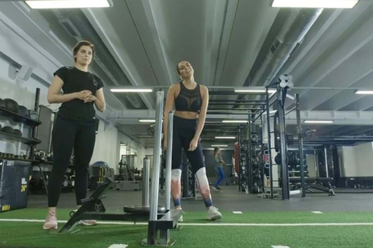 Two women standing on artificial turf in a gym with exercise equipment around them