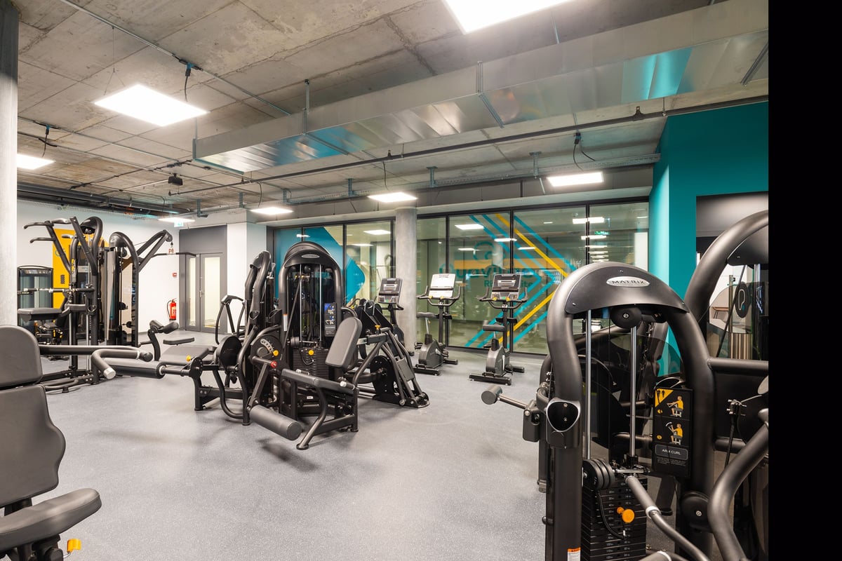PureGym Genf Le Bouchet: Gym with weight machines, free weights, benches, mirrors, and teal and yellow accent walls.