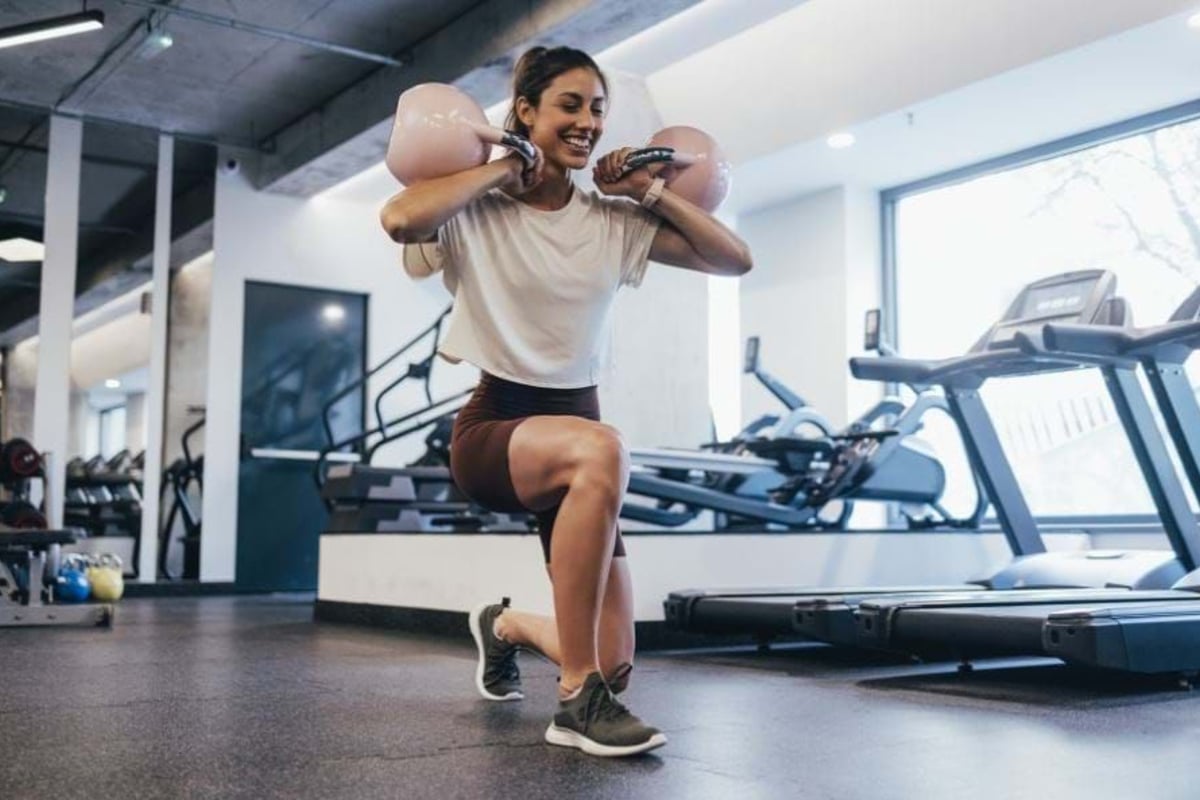 Woman doing lunges with kettlebells on shoulders in gym with treadmills nearby