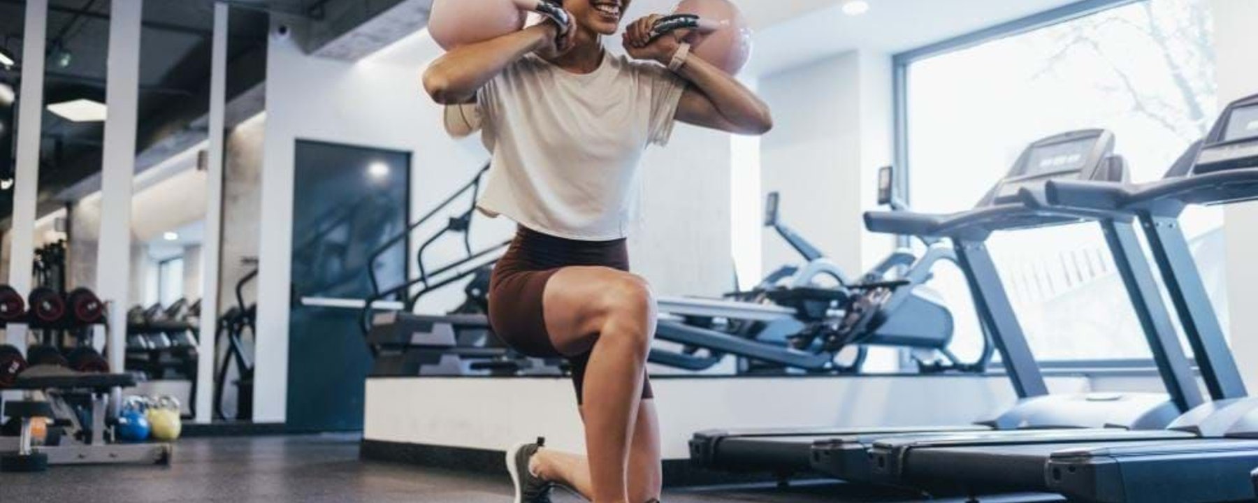 Woman doing lunges with kettlebells on shoulders in gym with treadmills nearby