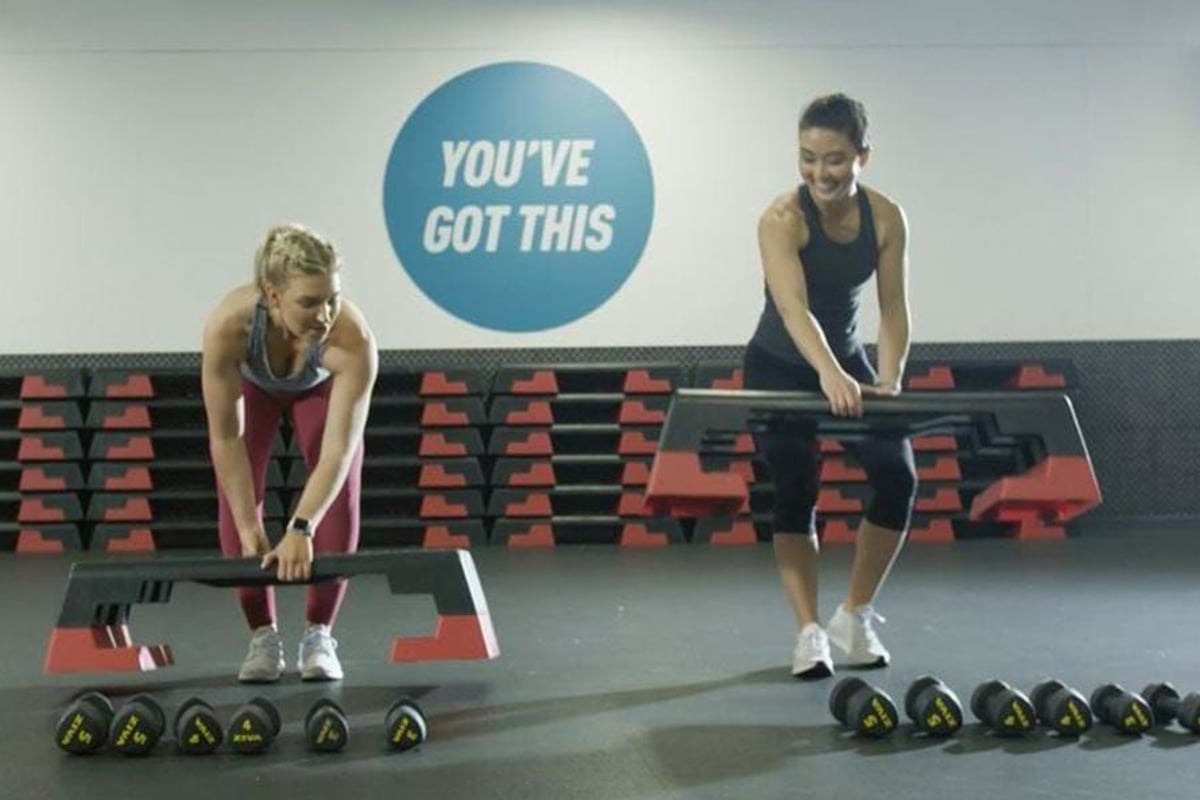 Two women lifting step platforms in a gym with dumbbells and a motivational sign