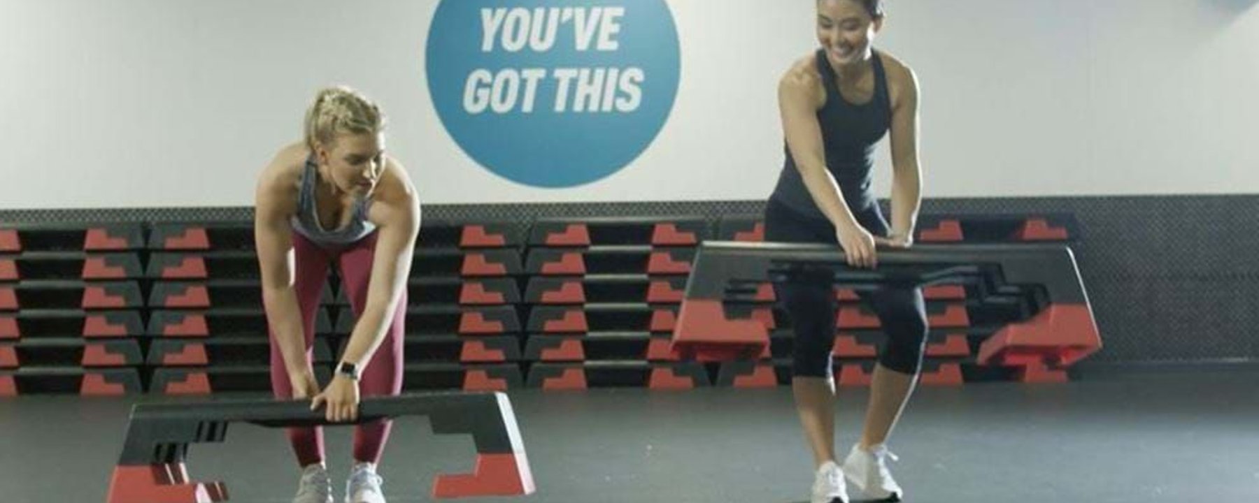 Two women lifting step platforms in a gym with dumbbells and a motivational sign