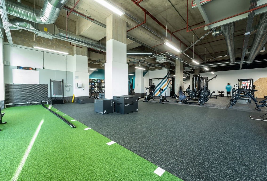 PureGym London Woolwich: Gym studio with green turf area, weight racks, exercise equipment, and rubberized flooring.