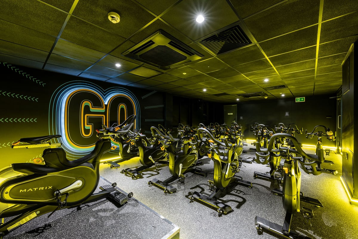 PureGym London Putney: Rows of stationary bikes, bright neon "GO" sign, dark interior with yellow accents.