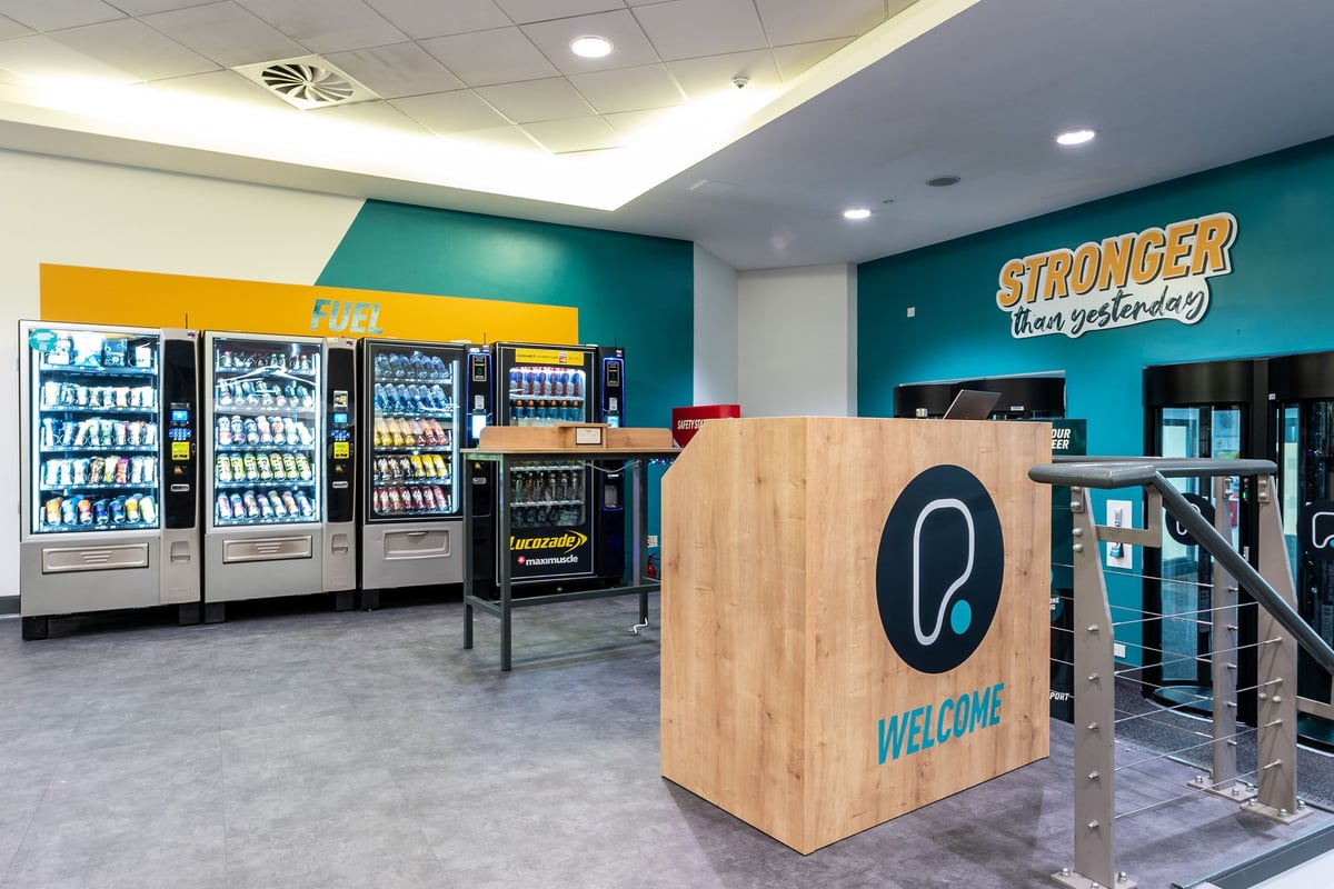 PureGym Cambridge Leisure Park: Vending machines, snack racks, and a welcome desk with "Stronger" signage.