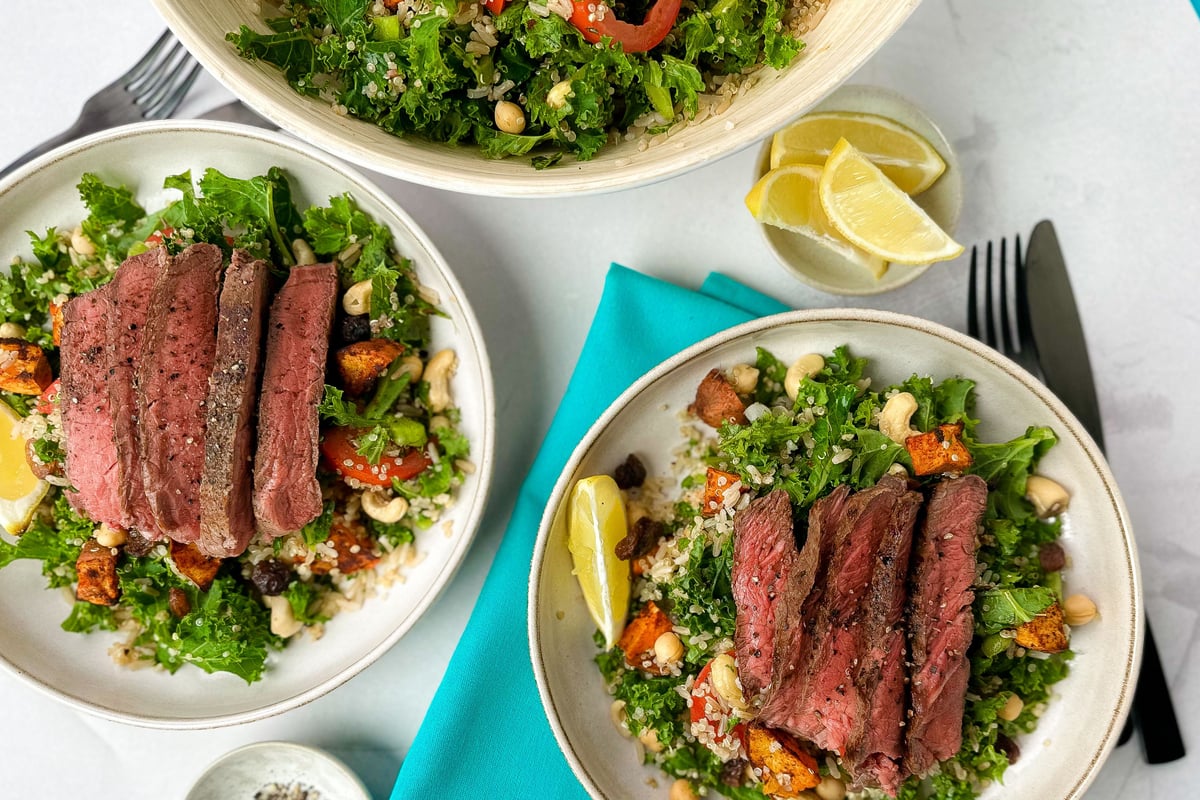 Three bowls of sliced steak on kale salad with roasted vegetables and cashews