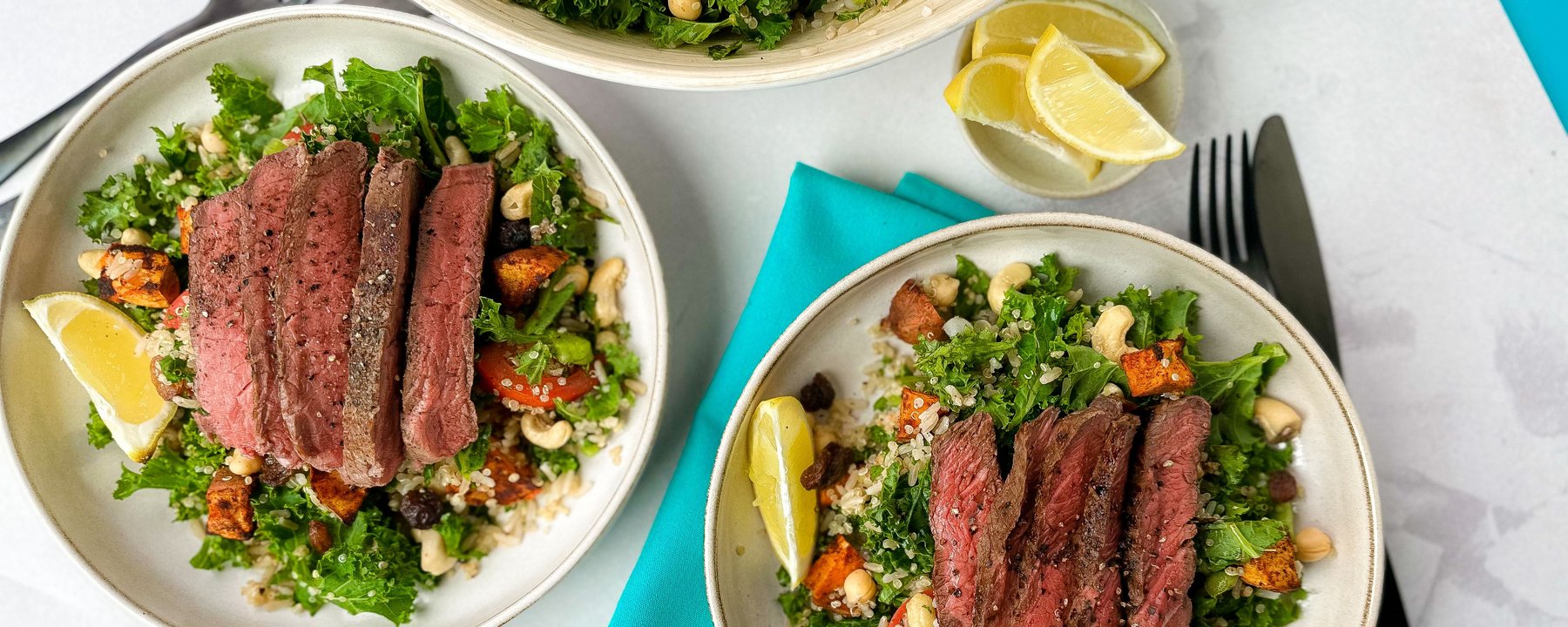 Three bowls of sliced steak on kale salad with roasted vegetables and cashews