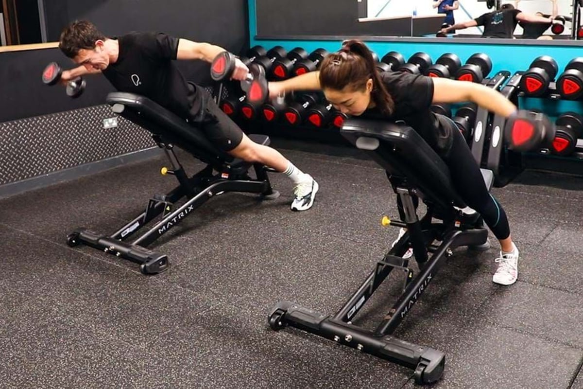 Two people lifting dumbbells while lying face down on inclined benches in a gym.