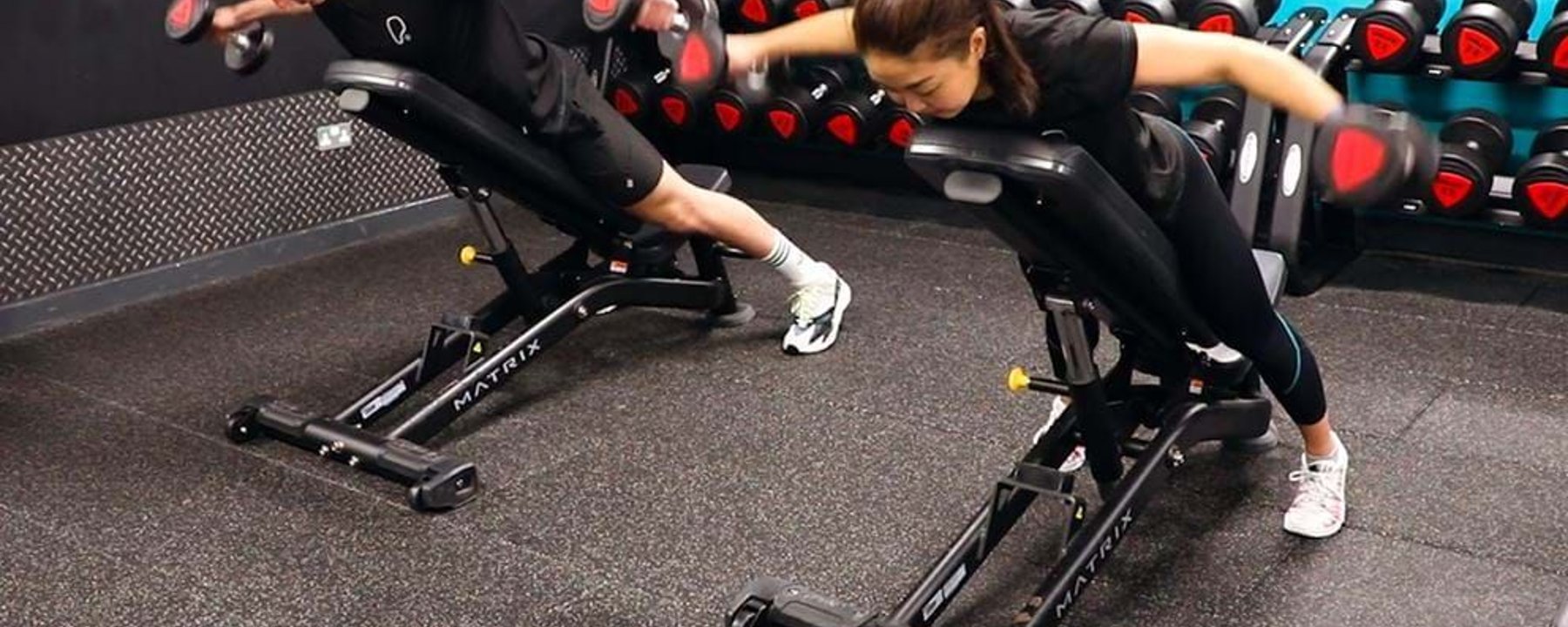 Two people lifting dumbbells while lying face down on inclined benches in a gym.