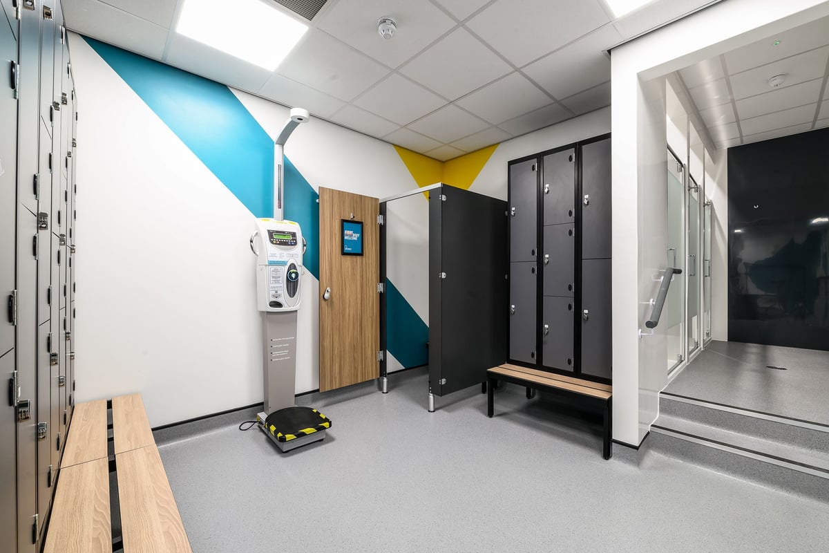 PureGym London Wimbledon: Locker room with black lockers, wooden benches, water fountain, and colorful wall accents.