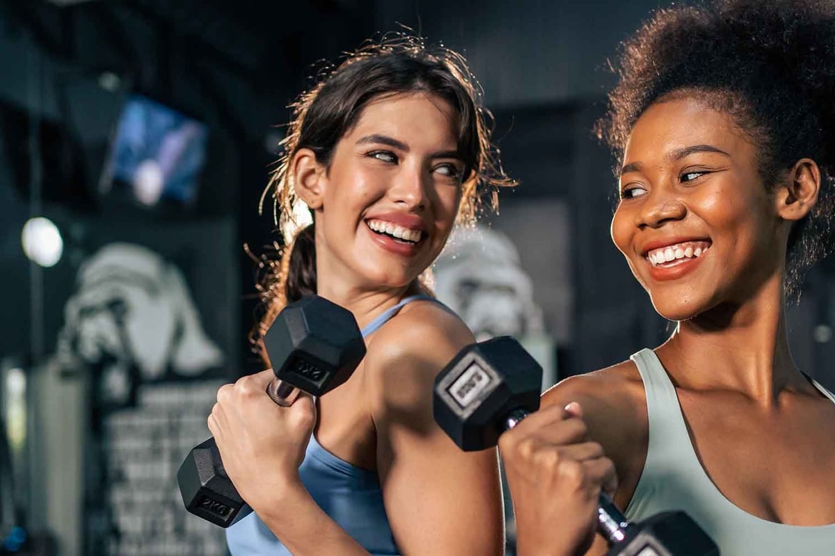 Zwei lächelnde Frauen trainieren mit Hanteln in einem modernen Fitnessstudio.