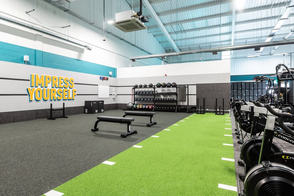 PureGym Winchester: Weight racks, benches, green turf area, equipment lined along mirrored wall.