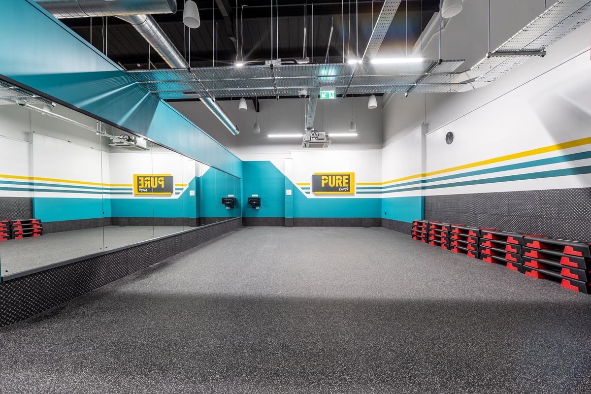 PureGym Dungannon: Rubberised flooring, red benches, speakers, and turquoise accent walls in a fitness studio.