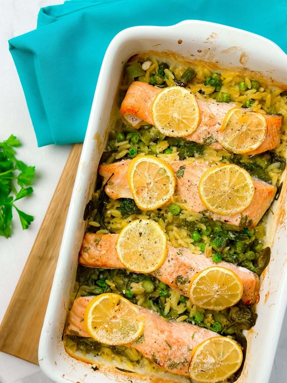Baked salmon fillets topped with lemon slices on a bed of rice and peas in a white dish