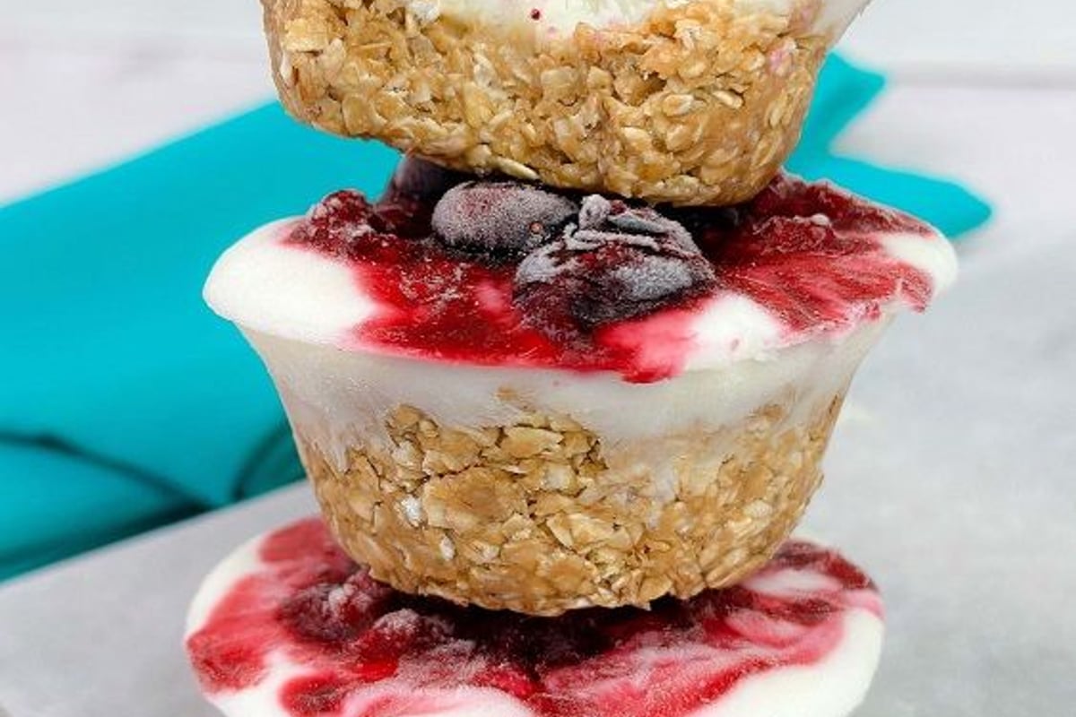 Stack of three frozen yoghurt and berry oat cups on a white surface with a blue cloth in b