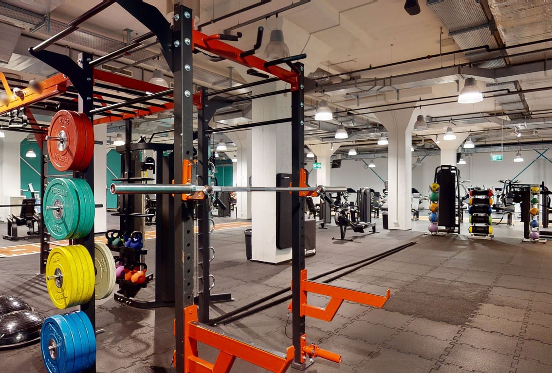 PureGym Basel Markthalle: Gewichtheberständer, Hantelscheiben, Kniebeugen, Bänke und gummierte Fussböden eines Fitnessraumes.