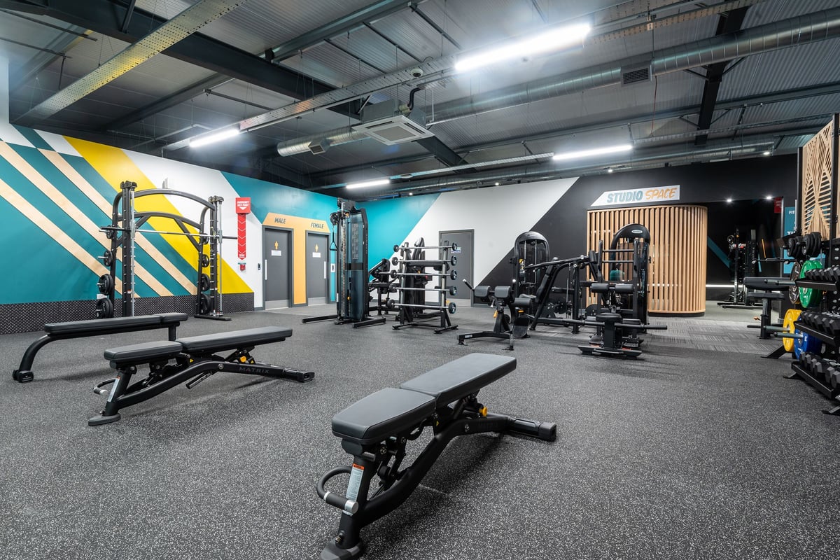 PureGym Glasgow Rutherglen: Gym studio with weight racks, benches, rubberized flooring, and colourful accent wall murals.