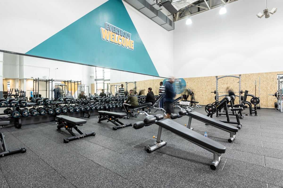 PureGym Rochdale: Rows of weight benches, free weights, squat racks, and weighted sleds in a gym studio.