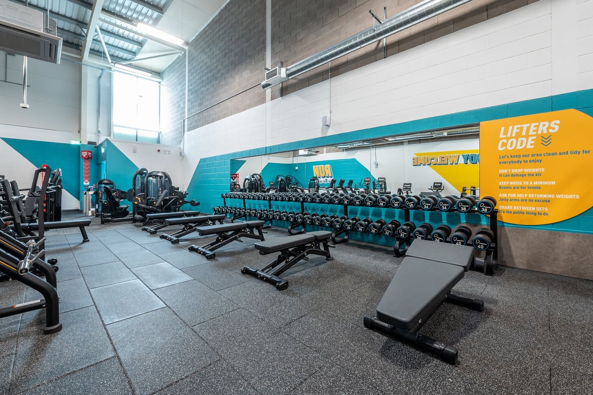 PureGym Shepton Mallet: Rows of teal weight benches, equipment racks with dumbbells, and rubberized gym flooring.