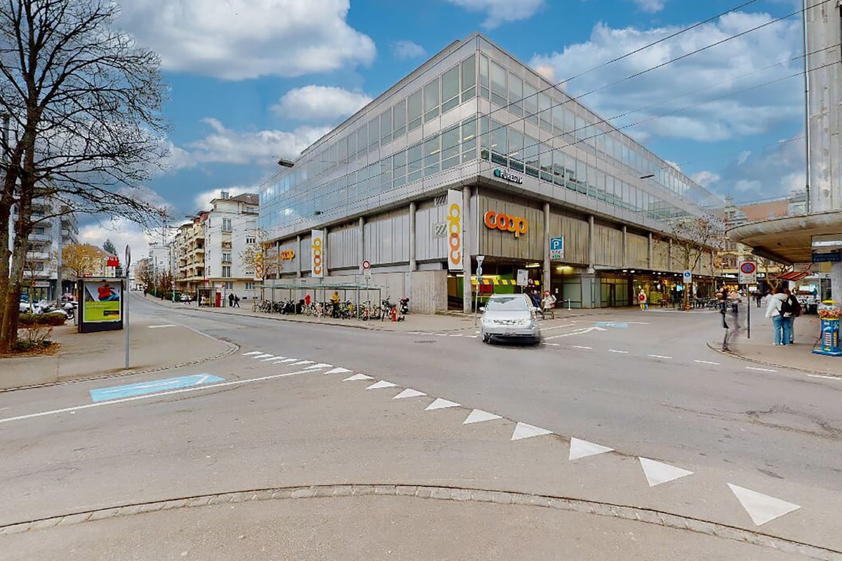 PureGym Biel Bienne: Une rue urbaine avec un bâtiment moderne à façade vitrée, des piétons et des véhicules en stationnement visibles.