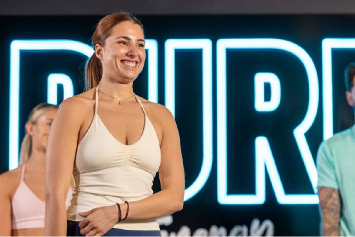 Frau im weißen Sportoberteil lächelnd vor blauem Neonschild mit anderen Personen daneben.