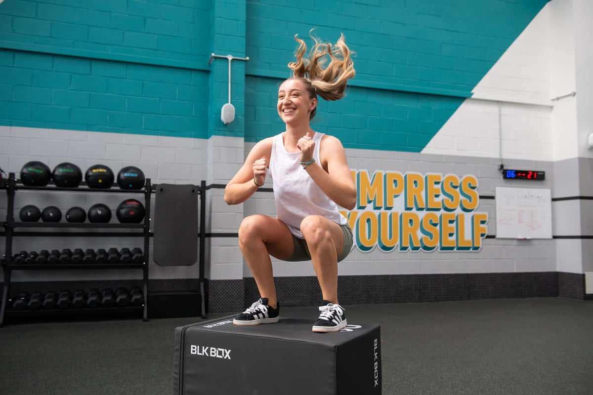 Woman jumping on a black exercise box in a gym with "Impress Yourself" on the wall behind