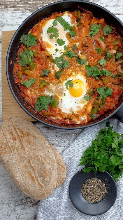 Two eggs cooked in tomato sauce with herbs in a pan, bread, parsley, and pepper bowl