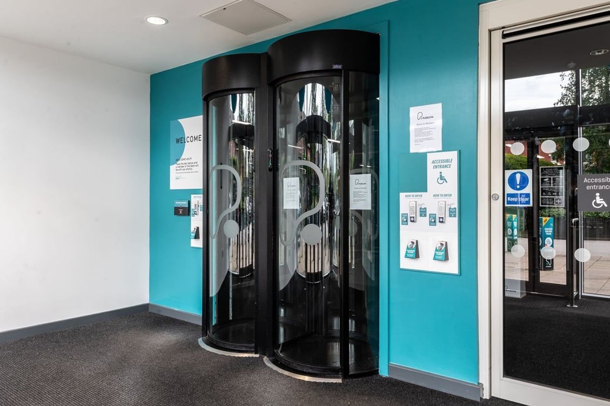PureGym Oldham: Two cylindrical revolving door machines in a teal-colored room interior.