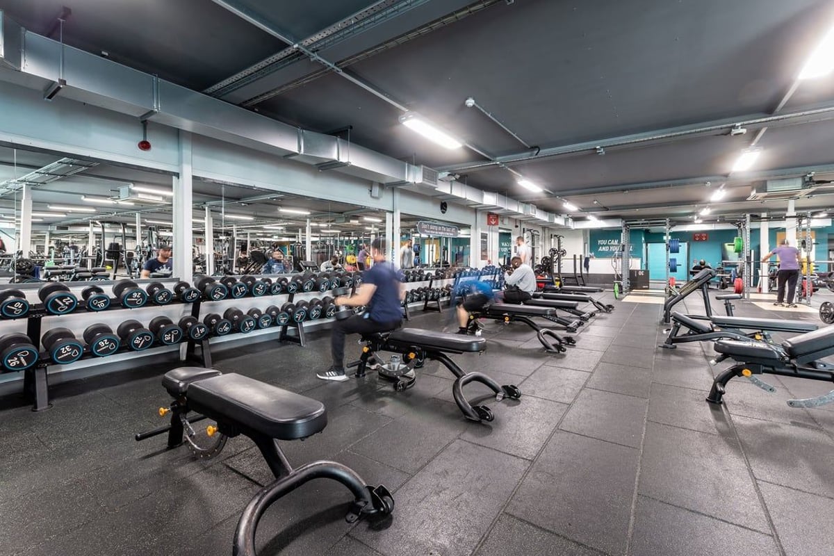 PureGym Oldham: Rows of free weights, benches, resistance machines, and rubberized flooring in a dimly lit gym interior.