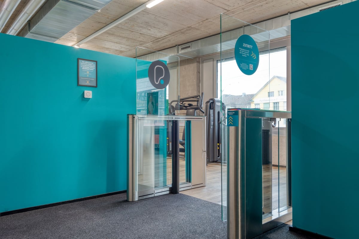 PureGym Lenzburg : portes d'accès au gymnase et zone de réception avec murs et signalisation bleu sarcelle.