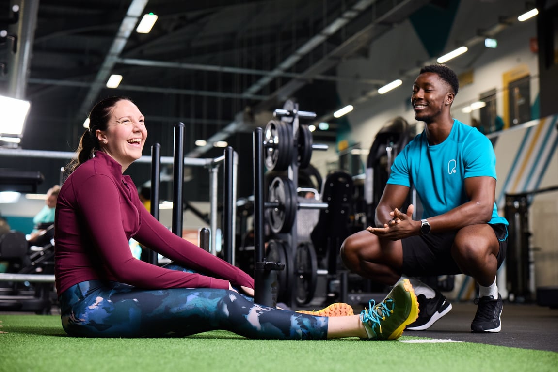 A man and woman sit on the gym floor, engaged in conversation, surrounded by exercise equipment.
