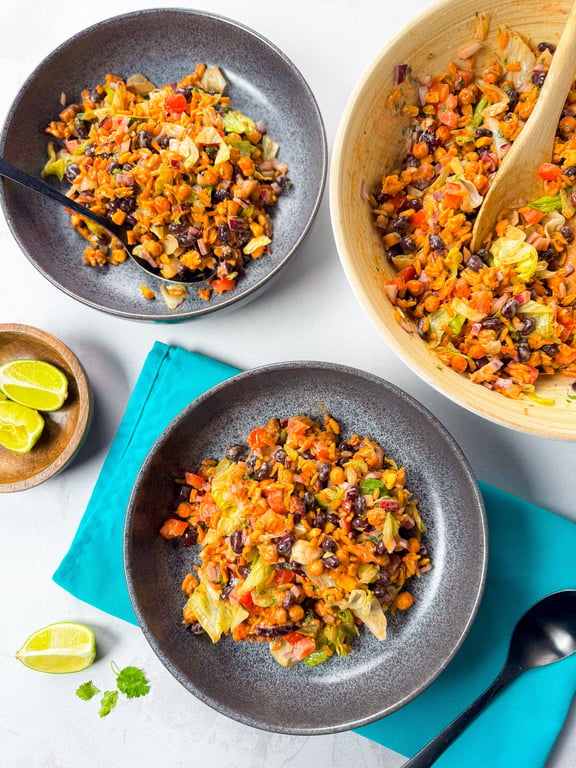 Two bowls and a wooden bowl filled with mixed bean and vegetable salad with lime wedges