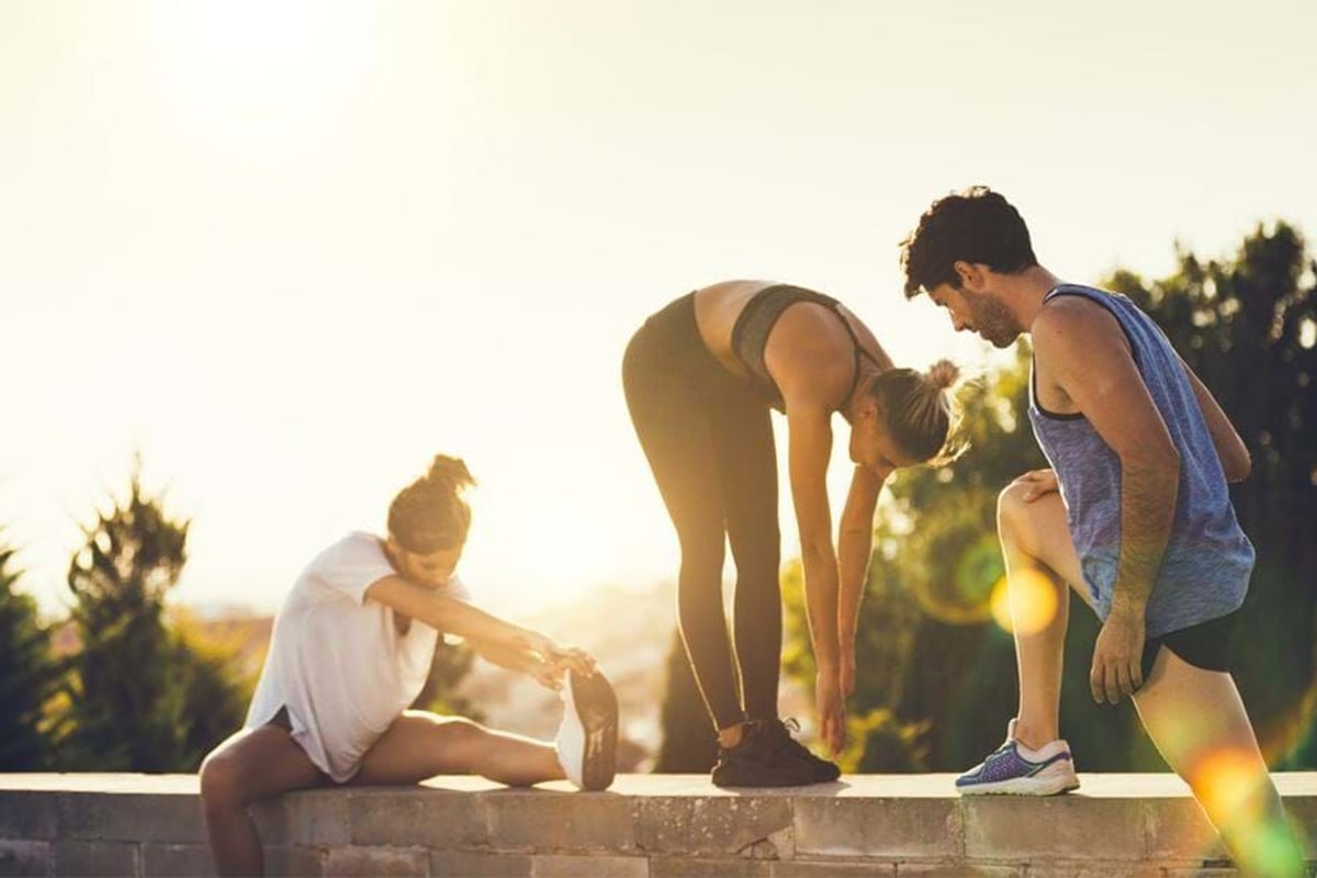 Three people stretching outdoors at sunset on a low wall with trees in background