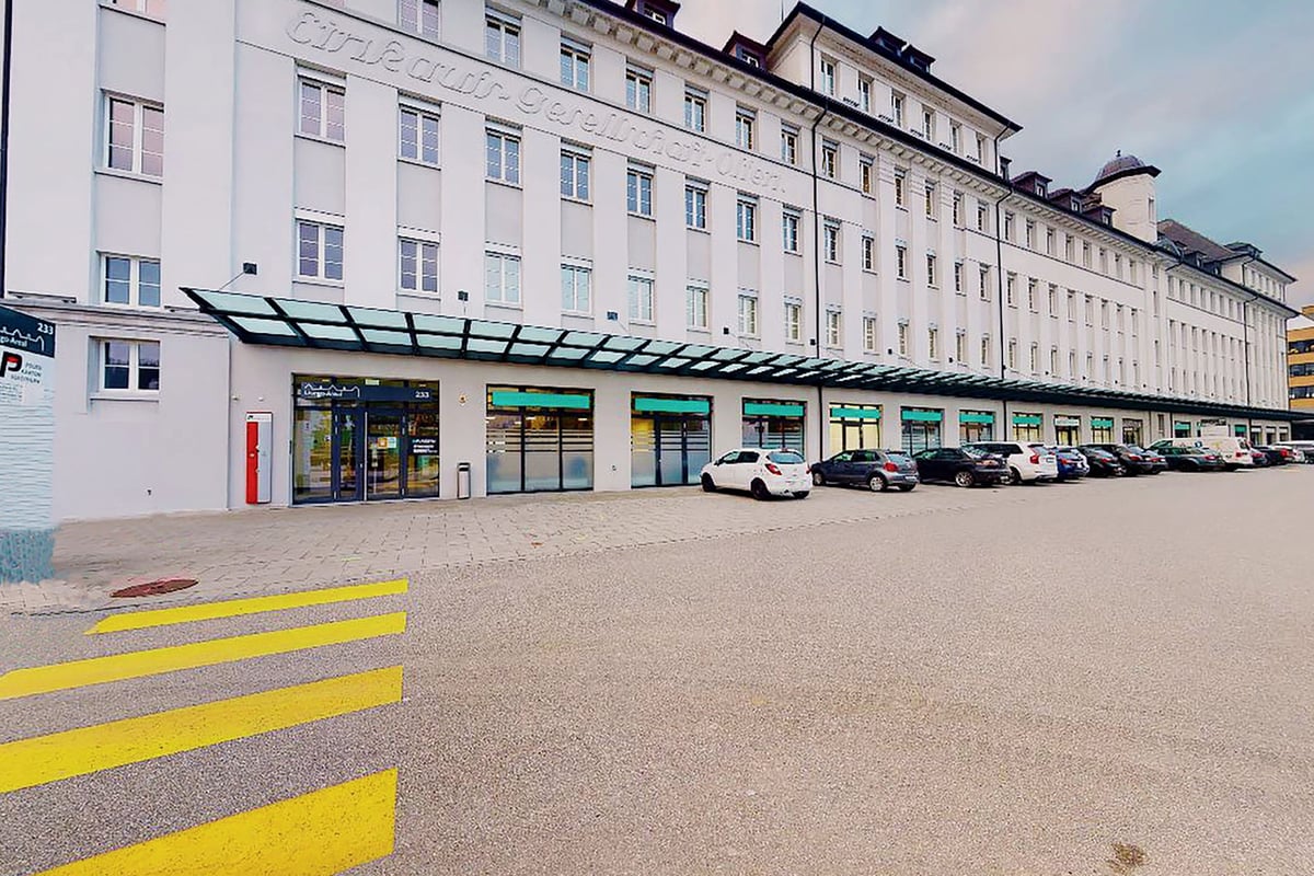 PureGym Olten: Aussenansicht des beigefarbenen Bürogebäudes mit Geschäften im Erdgeschoss, geparkten Autos und PUREGYM-Eingang.