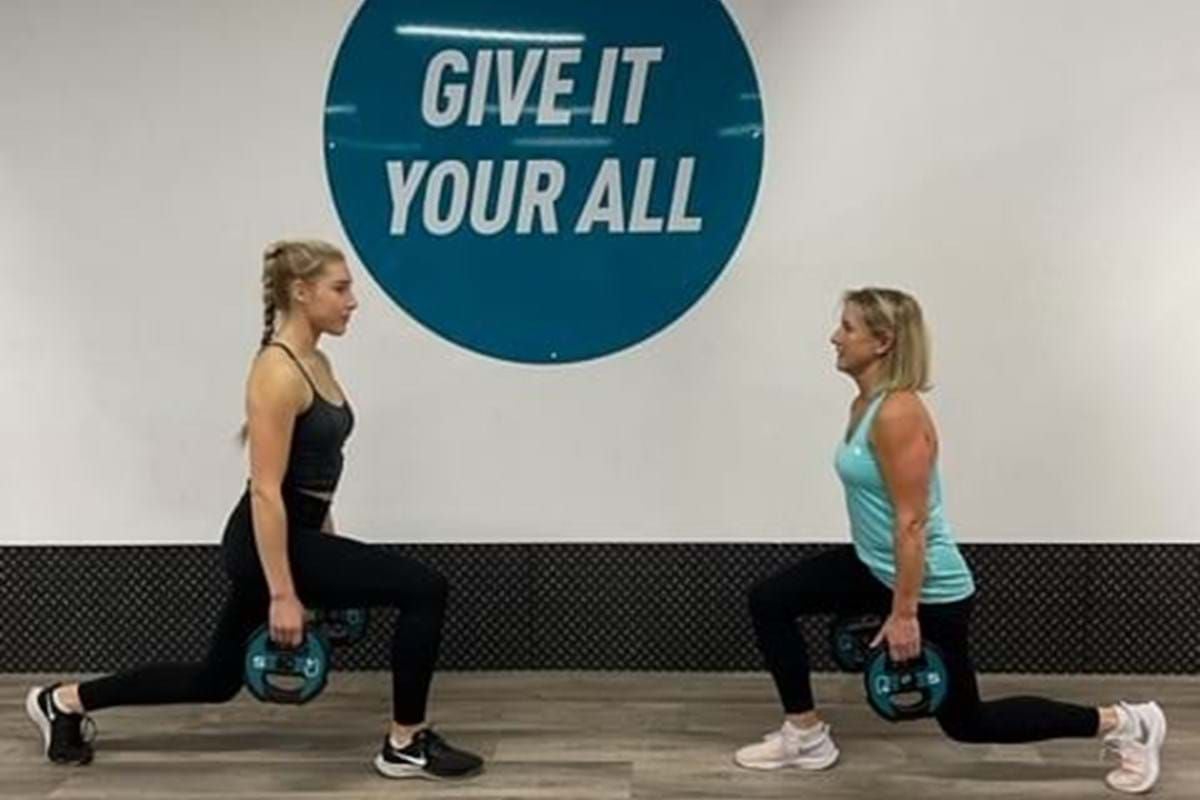 Two women doing lunges with weights in front of a wall sign saying Give it your all