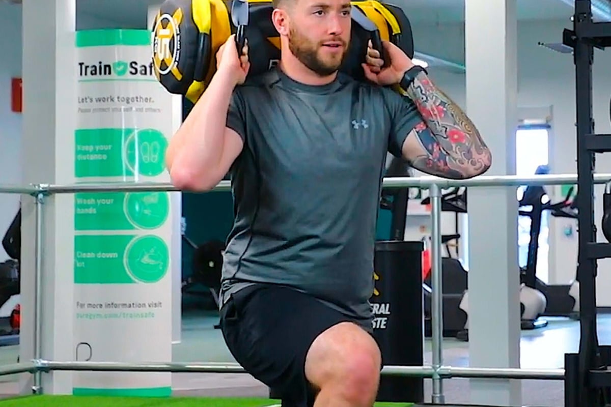 Man with tattooed arm doing weighted lunges in gym with safety sign behind him
