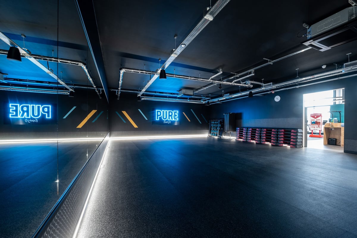 PureGym Trowbridge: Fitness studio with teal lighting, mirrored walls, and free weights area.