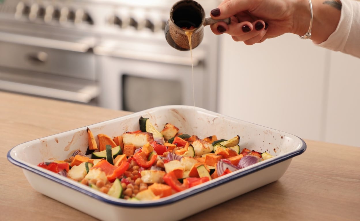 Hand drizzling sauce over roasted vegetables and halloumi in white enamel baking tray