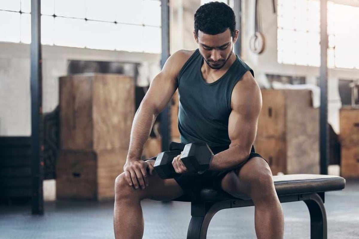 Man in black tank top lifting dumbbell while sitting on gym bench