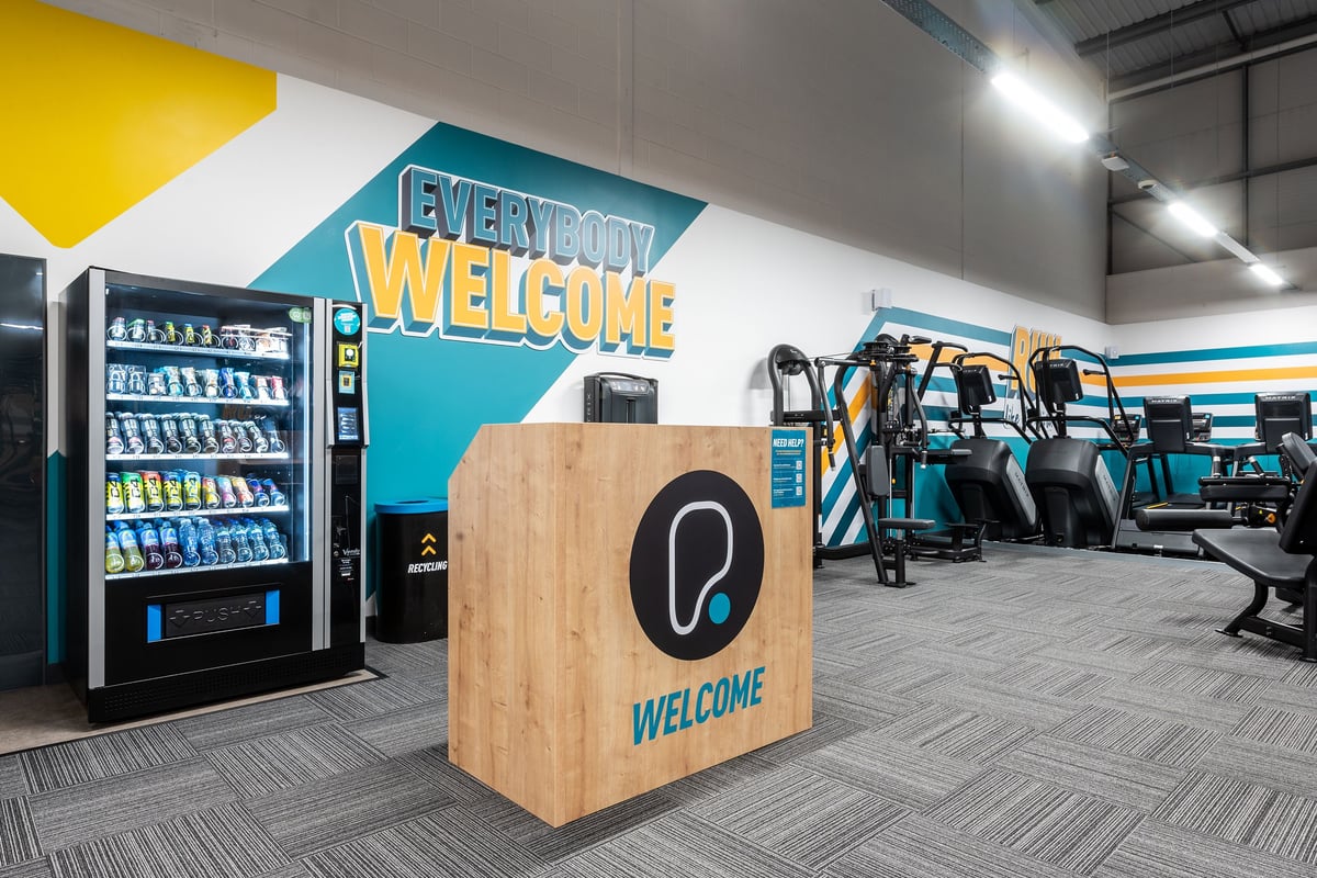 PureGym Harwich: Rows of exercise machines, vending unit, welcome desk in teal and yellow decor.